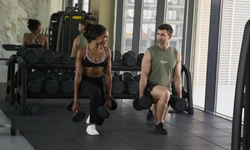 Personal trainer Rozzie Kinyua and a male client performing dumbbell lunges together in a Dubai gym.