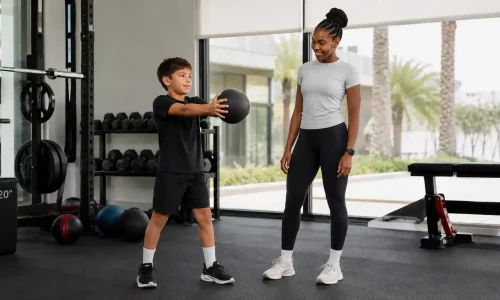 Young boy doing a medicine ball exercise with a female trainer during a supervised kids strength training session in Dubai.