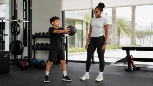 Young boy doing a medicine ball exercise with a female trainer during a supervised kids strength training session in Dubai.
